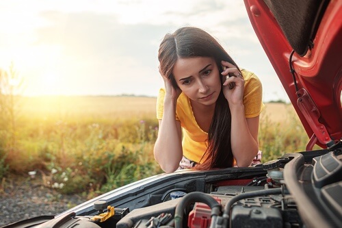 Cha-Ching! 5 Ways to Save on Domestic Vehicle Repair with John's Auto Pros in Escondido, CA; image of young woman stranded on the side of sunny desert road with hood of car raised while leaning over the car with hand on the back of her head while on the phone