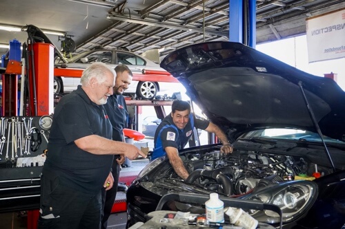 Auto Repair in Escondido CA with John's Auto Pros; image of owner John Stacey, Service Advisor and certified ASE tech pictured standing over black car in shop, engine while talking it over