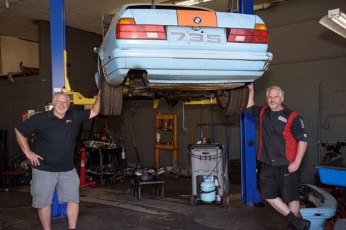 Racing car service and repair in Escondido, CA image of John's Auto Pros owner John Stacey and ASE certified tech Dan pose next to light blue BMW race car on lift in air in shop bay